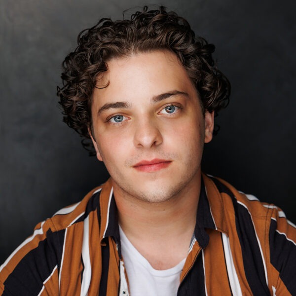 A young person with short curly brown hair and blue eyes, wearing a brown, black, and white striped shirt over a white T-shirt, looks directly at the camera against a dark background.