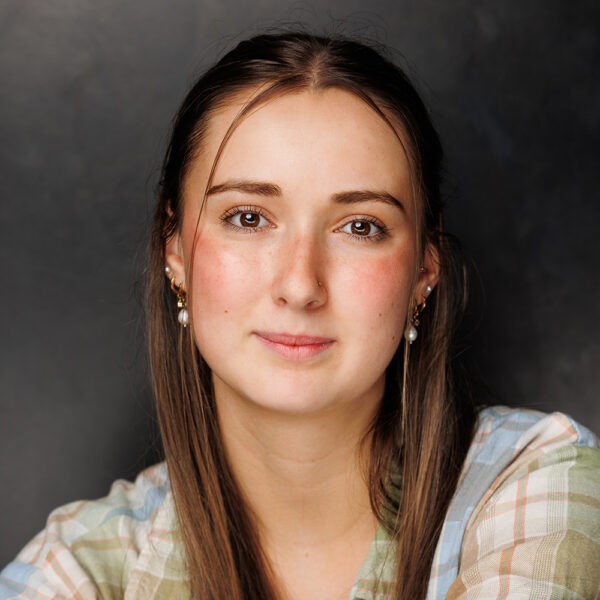 A young woman with straight brown hair and fair skin looks at the camera with a slight smile, wearing a checked shirt and pearl earrings, against a dark, plain background.