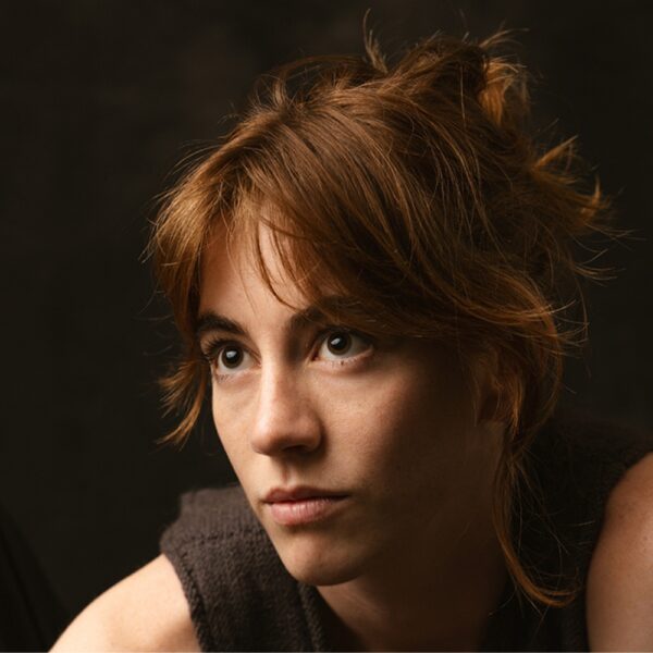 A young person with tousled, reddish-brown hair gazes thoughtfully upwards. The background is dark, and soft lighting highlights their face, showing a serious, contemplative expression.