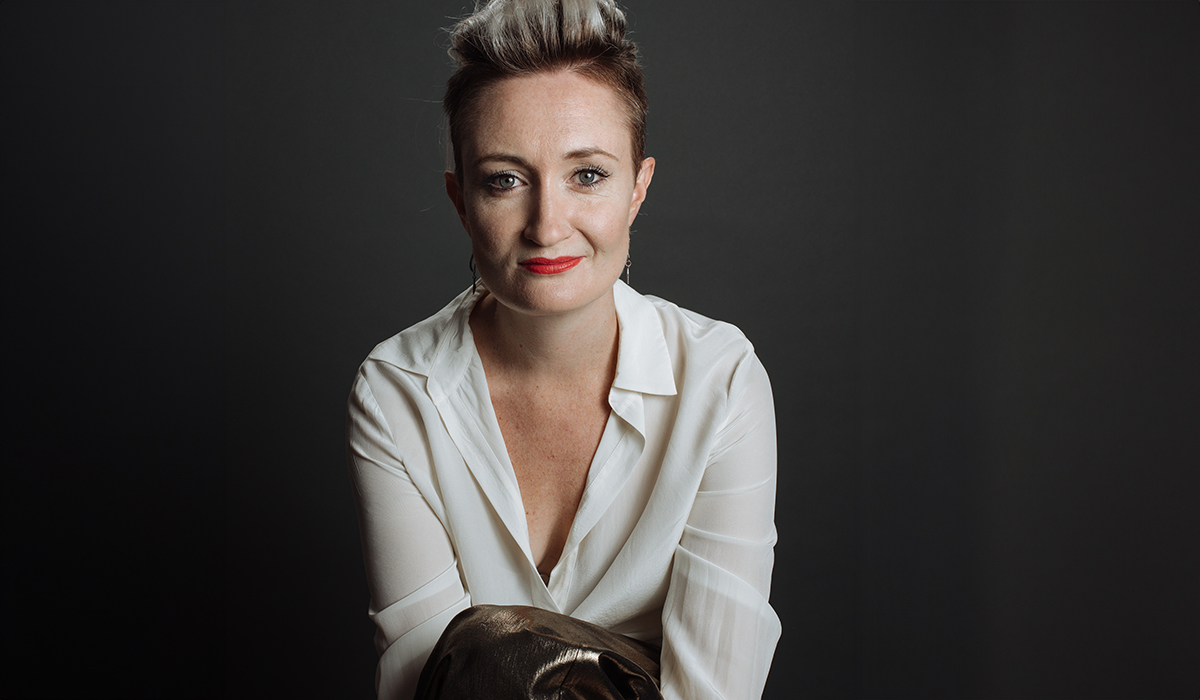 A person with short, styled fair hair and red lipstick sits with arms crossed, wearing a white blouse and dark trousers, posing against a dark, plain background.