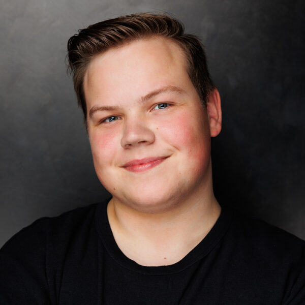 A young person with short brown hair and blue eyes smiles at the camera, wearing a black shirt against a dark, plain background.