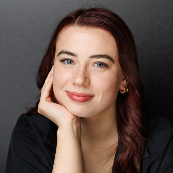 A woman with long auburn hair and blue eyes smiles gently, resting her chin on her hand. She wears a black top, small gold hoop earrings, and sits in front of a plain dark background.