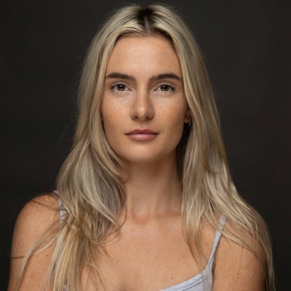 A woman with long blonde hair and light skin looks directly at the camera with a neutral expression, wearing a light-coloured vest top against a dark background.