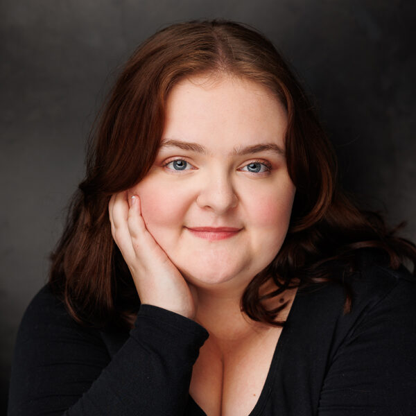 A woman with fair skin and brown hair smiles gently, resting her head on her hand. She is wearing a black top and is posed in front of a dark, blurred background.