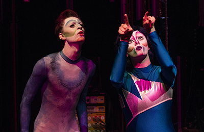 Two performers in colourful, dramatic make-up and tight costumes stand under stage lighting. One looks upward, while the other points ahead, both with expressive faces, suggesting a theatrical scene or circus performance.