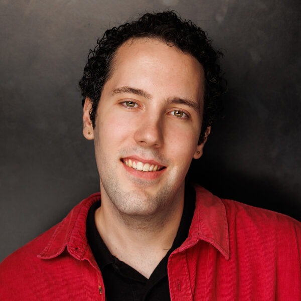 A young adult with curly dark hair and light skin smiles at the camera. They are wearing a red collared shirt over a black top and are posed in front of a dark, plain background.