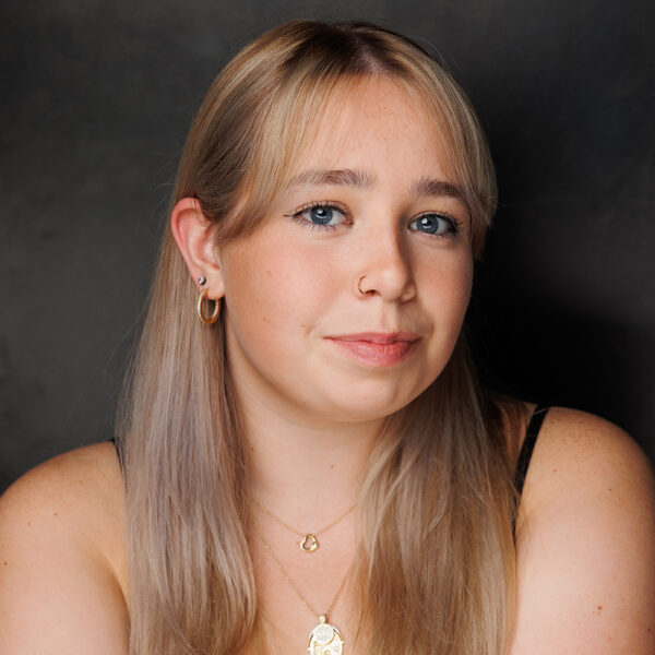 A young woman with long, straight blonde hair and blue eyes poses against a dark background. She wears gold hoop earrings, layered necklaces, and a black top, and has a small nose ring and a gentle expression.