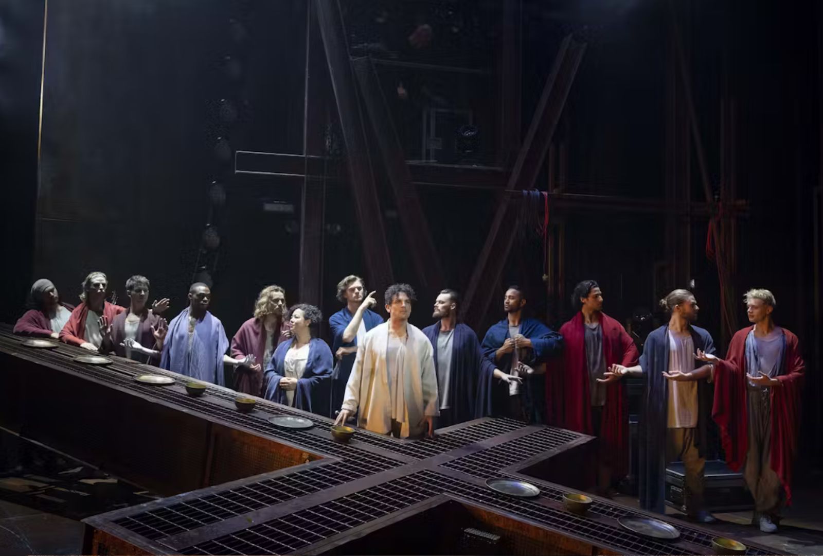 Michael Paynter with ensemble cast in Jesus Christ Superstar at the last supper. Plates and bowls are placed on an enormous cross in front of the actors.
