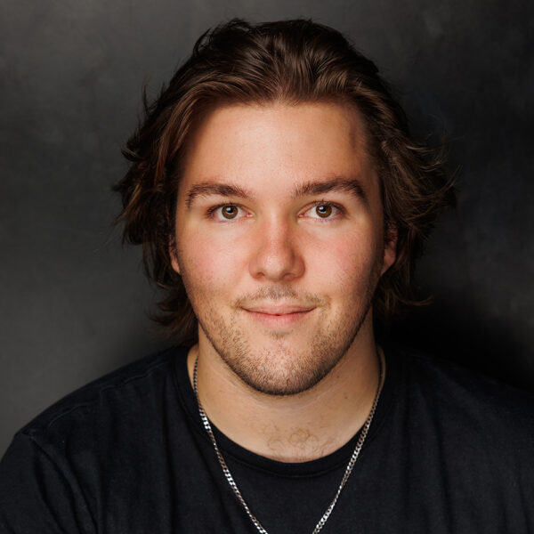 A young man with medium-length brown hair and a short beard smiles softly at the camera. He is wearing a black shirt and a silver chain necklace, with a dark, neutral background behind him.