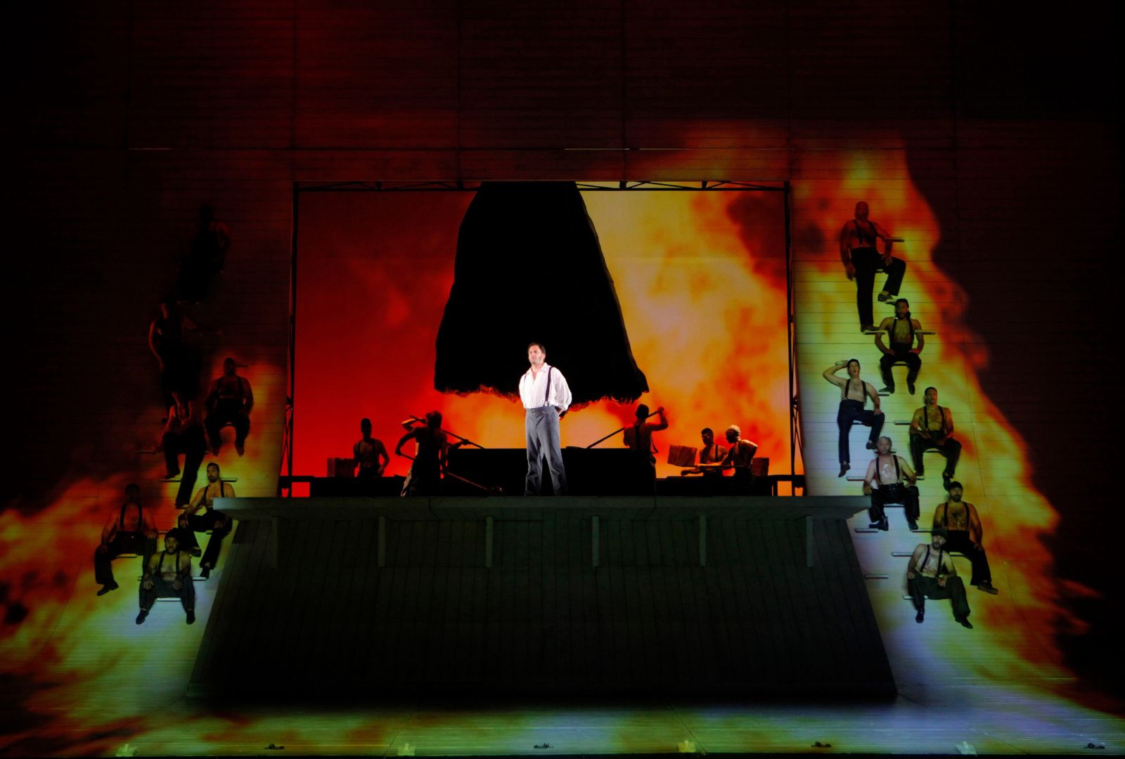 Production of Moby Dick. A man stands suspended on a stage in front of a fiery lit background with men either side of him watching on.