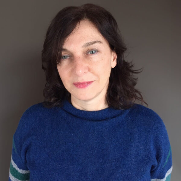 Woman with shoulder-length dark brown hair and blue eyes, wearing a blue jumper with green and white stripes on the sleeves, standing in front of a plain grey background, looking at the camera.