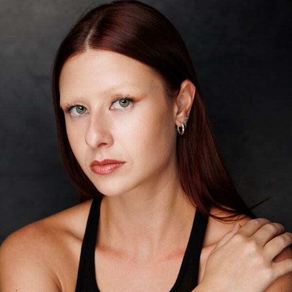 A woman with straight auburn hair, light skin, and green eyes looks into the camera. She wears a black sleeveless top, silver hoop earrings, and touches her shoulder, posing against a dark background.