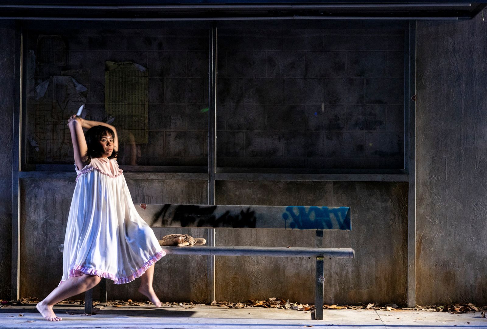 A young girl in a white nightgown strikes a dramatic pose on a bench at a dimly lit, graffiti-marked bus stop with scattered leaves on the ground.