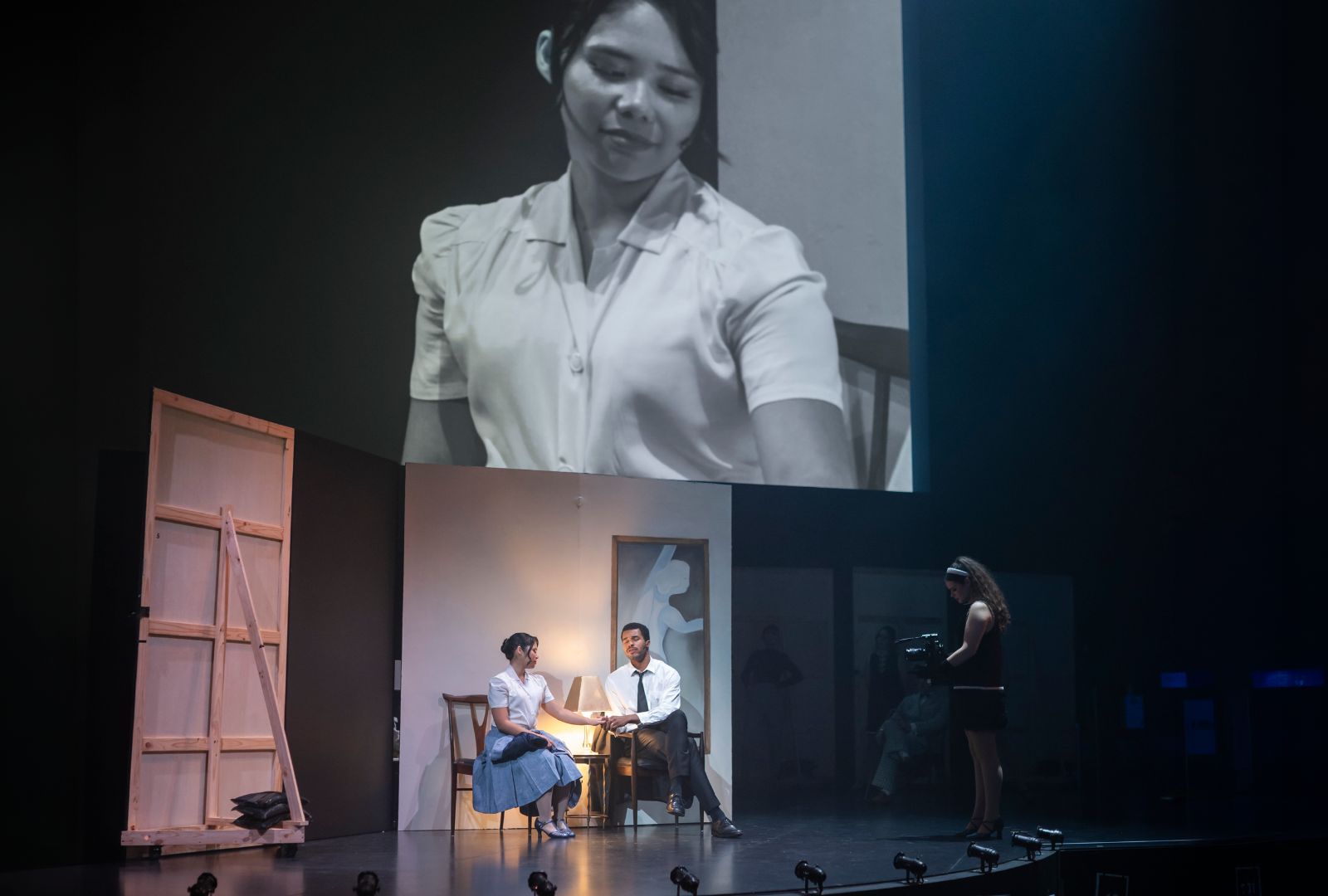 Two actors in period clothing seated on a stage with a camera person in front of them shooting an image projected onto a screen overhead.