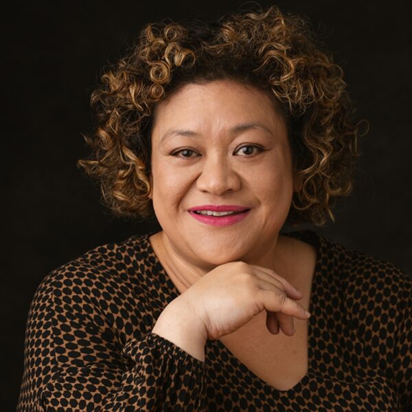 A woman with curly hair smiles, resting her chin on her hand. She wears a patterned brown and black top and has bright pink lipstick, against a dark background.