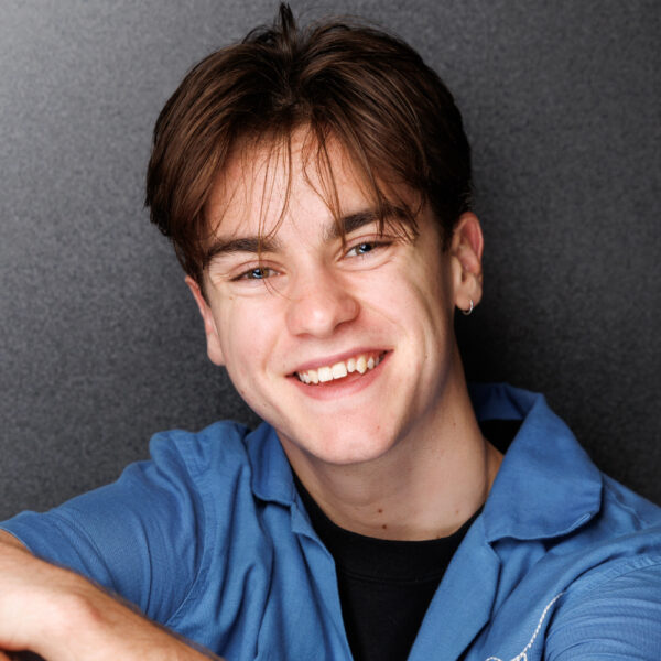 A young person with short brown hair and an earring smiles at the camera, wearing a blue shirt over a black top, against a dark grey background.