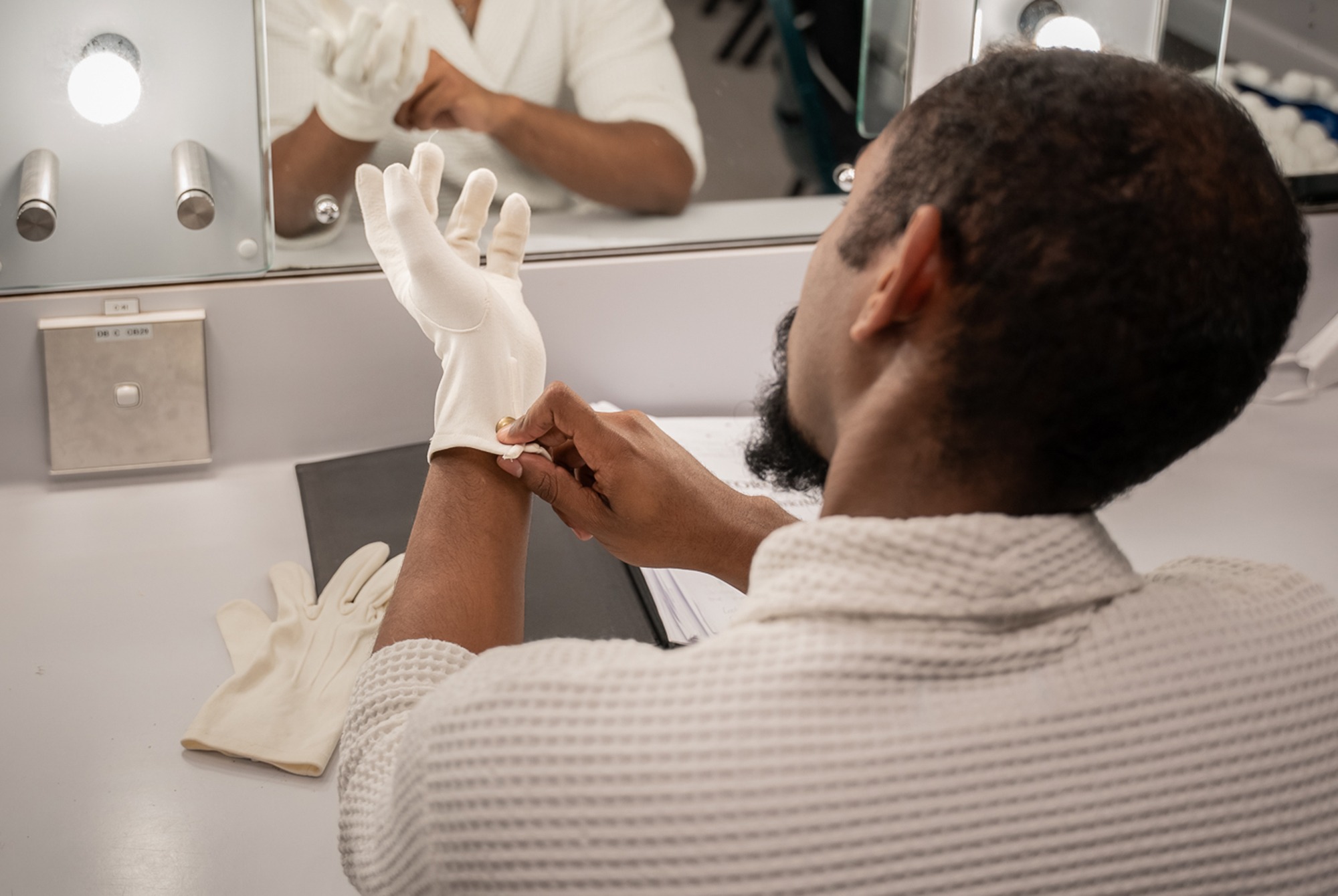 A person wearing a white dressing gown sits at a dressing table and puts on a white glove, with another glove, papers, and a mirror with lights visible on the table.