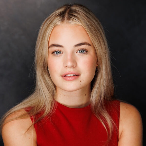 A young woman with long blonde hair and blue eyes, wearing a sleeveless red top, poses in front of a dark, neutral background. She is looking directly at the camera with a soft, relaxed expression.