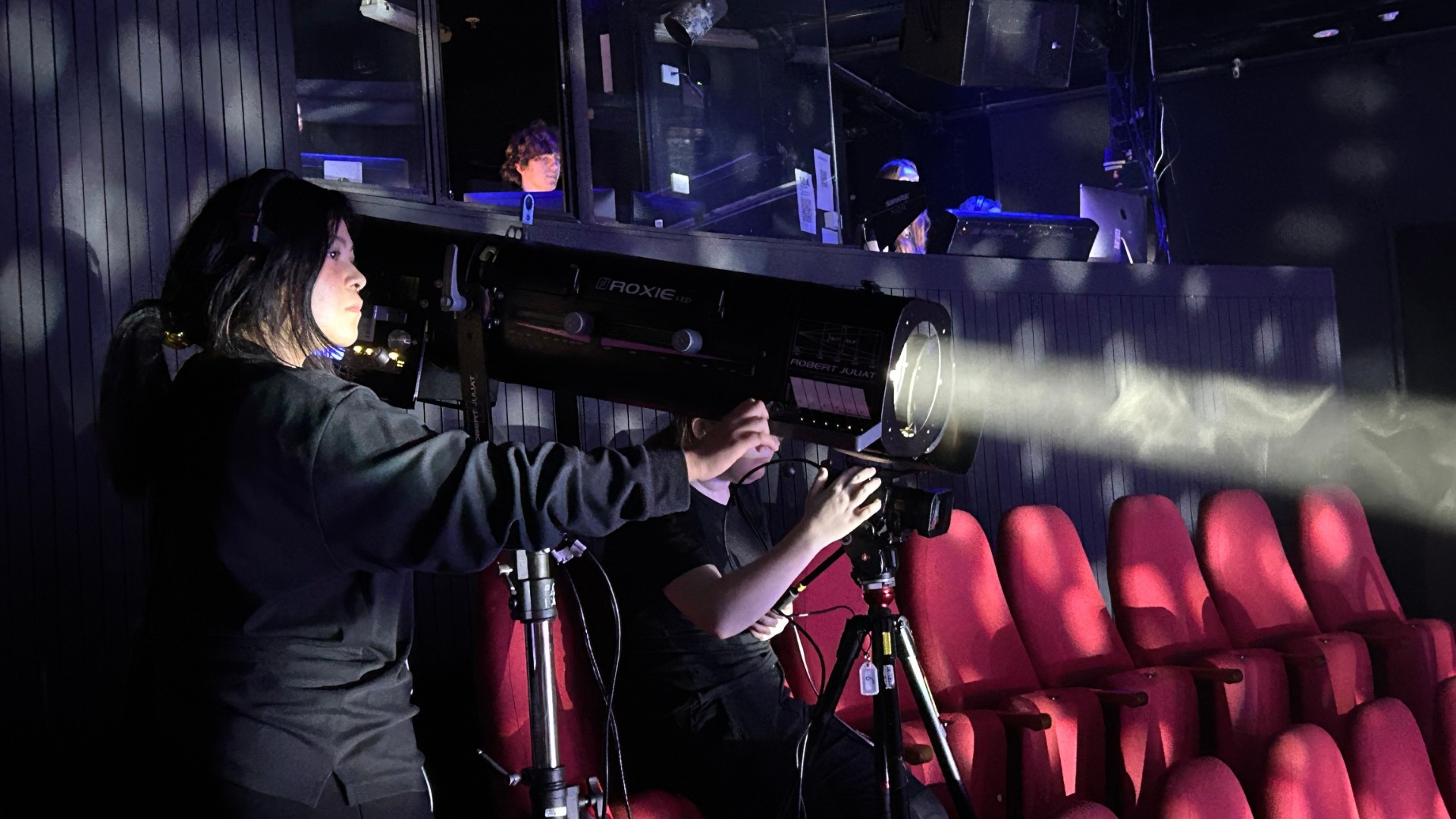 A person operates a spotlight in a dark theatre, directing a beam of light towards the stage. Another person sits beside them, and people are visible in a control box in the background. Red seats are in the foreground.