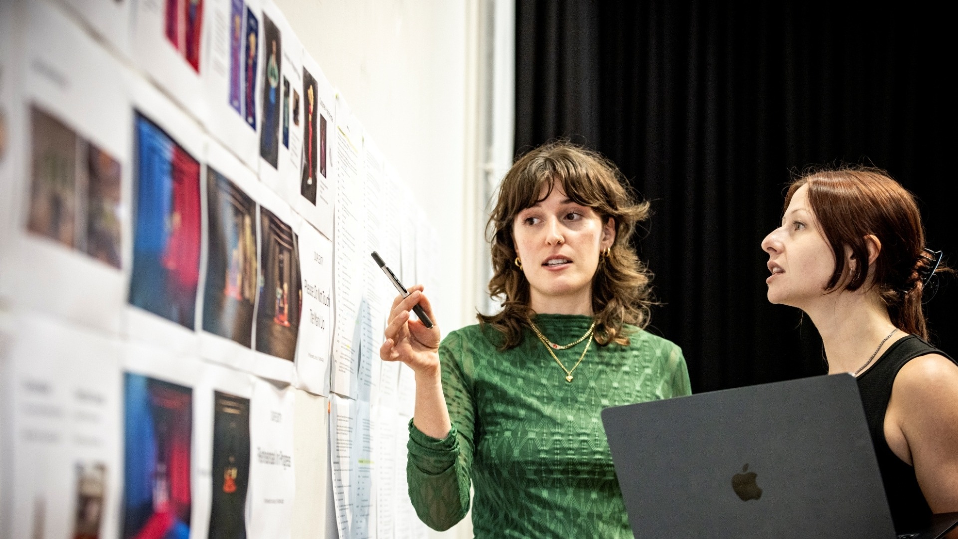 Two women stand by a wall covered with images and notes. One woman in a green top gestures at the wall with a pen, whilst the other, holding a laptop, looks on attentively.