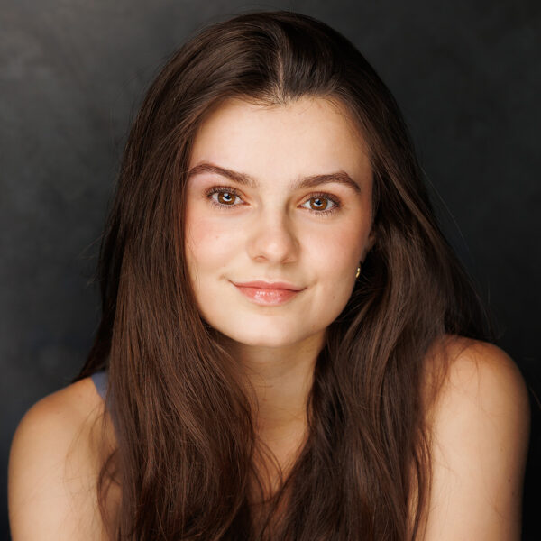 A young woman with long brown hair and light skin smiles softly at the camera against a dark, blurred background. She is wearing a sleeveless top and has natural make-up.