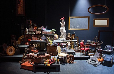 A theatrical stage filled with assorted vintage furniture, suitcases, flowers, and decorative objects, including a white statue, empty picture frames, and a rocking horse, all arranged in a cluttered, artistic display.