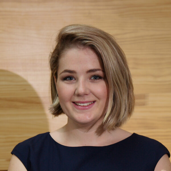 A woman with short blonde hair and blue eyes, wearing a navy blue top, smiles at the camera in front of a wooden background.
