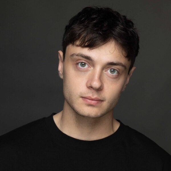A young man with short dark hair and light eyes is facing the camera with a neutral expression, wearing a black shirt against a dark, plain background.