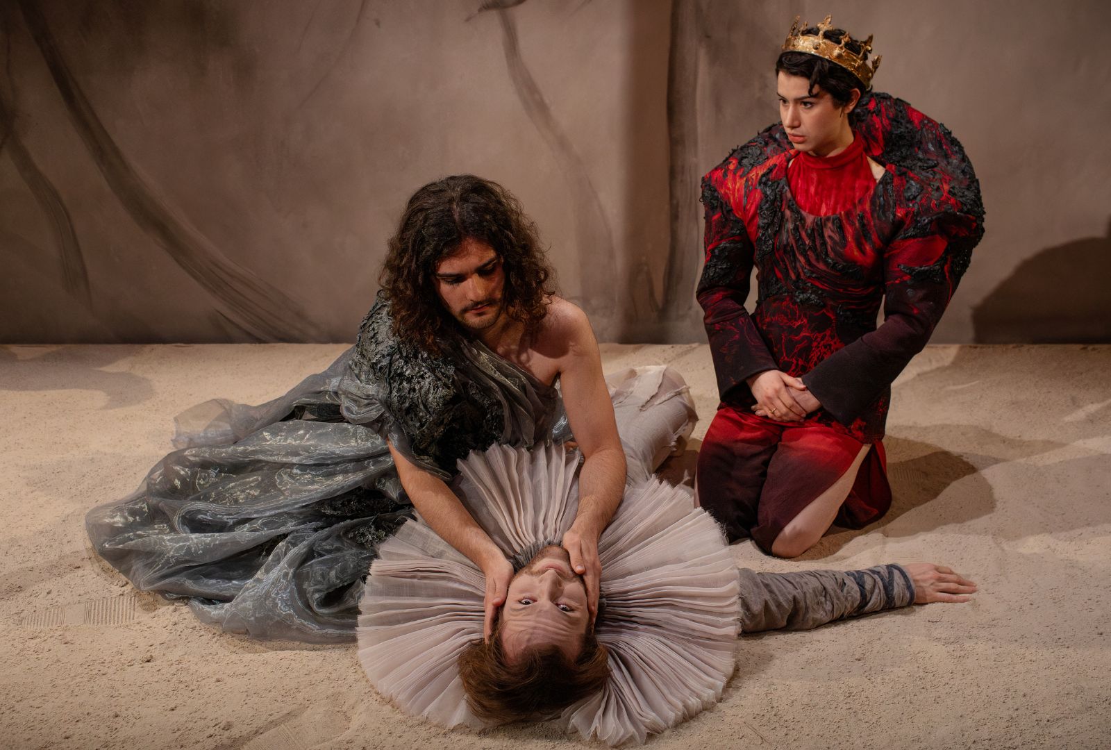 An actor wearing a loose, grey dress holds the face of a man on the ground. He has a large Elizabethan ruffle and lays on sand. Another actor sits on the right in a red and black avant-garde suit and crown.