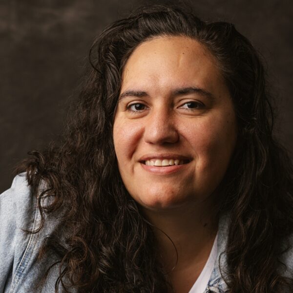 A person with long, curly dark hair and a light blue denim jacket smiles at the camera against a dark, neutral background.