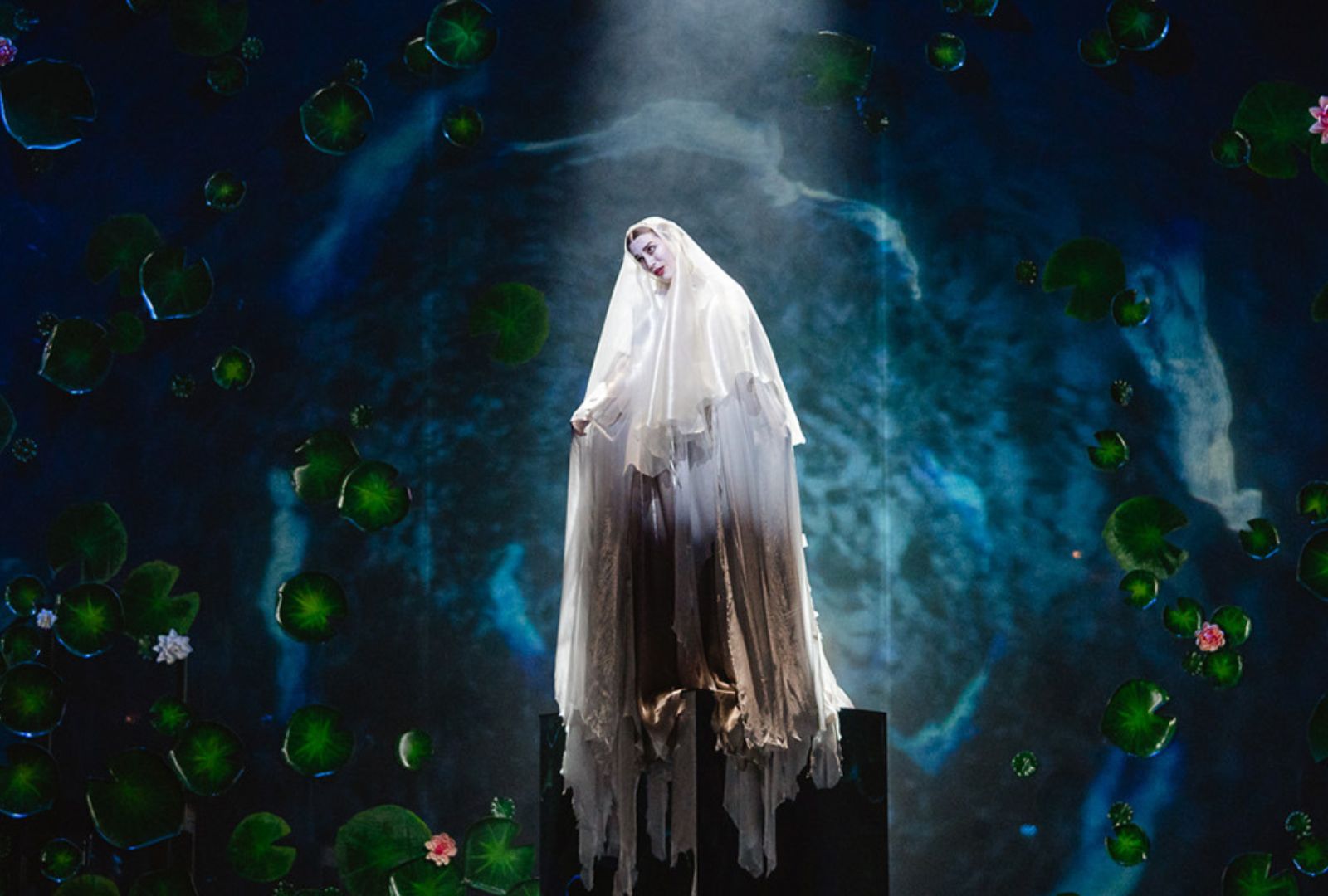 A performer dressed in white, draped clothing and wearing white face paint stands with water and lilies projected behind her. 