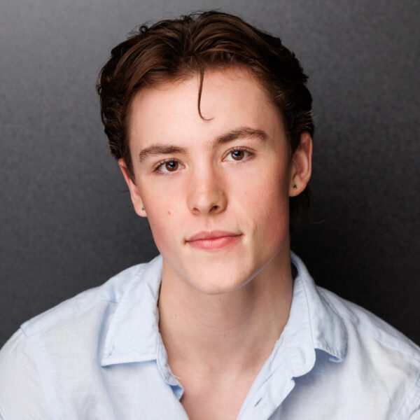 A young person with short brown hair and fair skin, wearing a light blue collared shirt, looks directly at the camera with a neutral expression against a plain dark background.