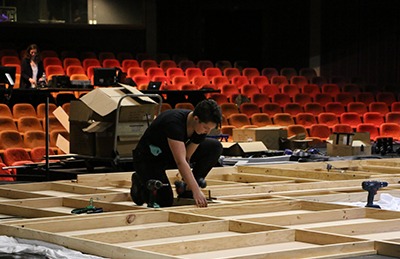 A person kneels on a wooden stage frame under construction, using tools, with scattered boxes and equipment about. Rows of empty red seats are visible in the background.