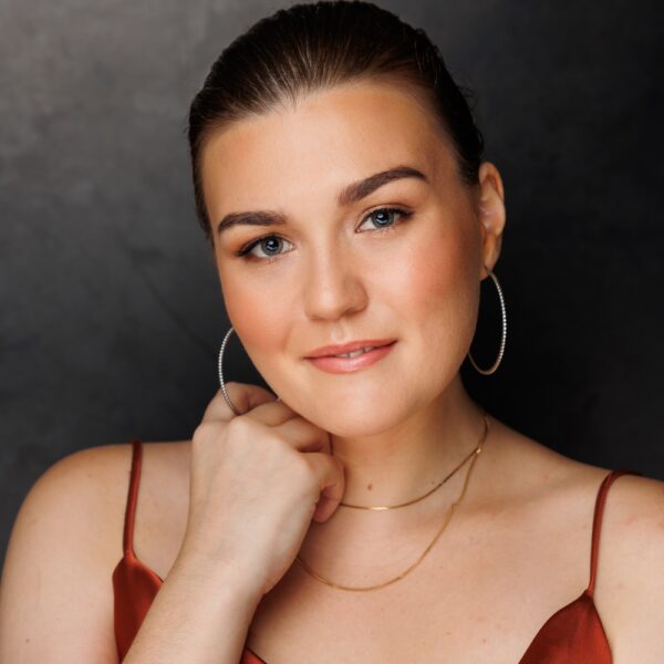 A woman with fair skin and brown hair, pulled back, wearing hoop earrings and layered gold necklaces, poses against a dark background. She is dressed in a satin brown top and smiles softly, with her hand touching her neck.