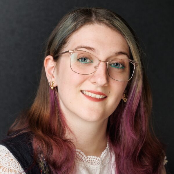 A young woman with glasses and long brown hair with purple highlights smiles at the camera. She is wearing a white lace top, gold earrings, and a dark waistcoat, with a dark, plain background.