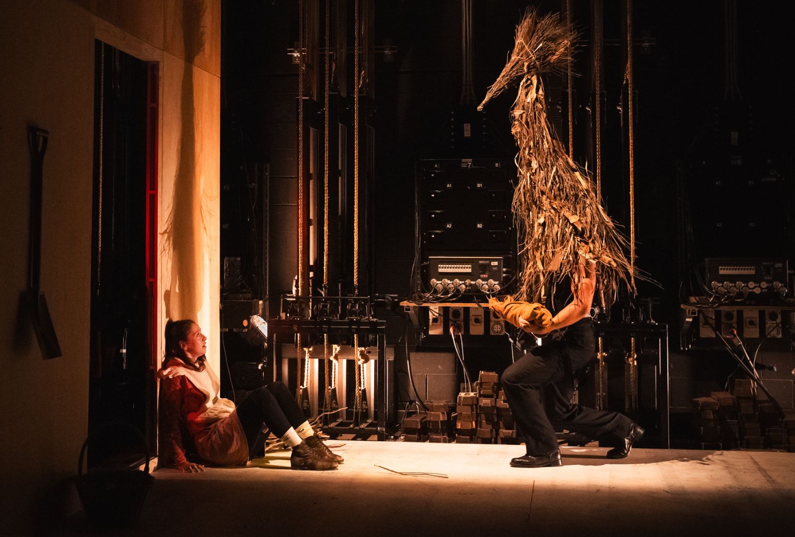 In a dimly lit theater, a person crouches against a wall with a fearful expression, while a performer in a tall, straw-covered costume with horns approaches them on stage. Stage equipment and cables are visible in the background.
