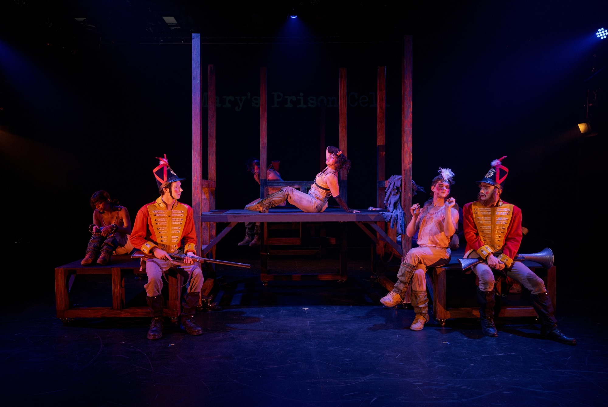 A theatrical stage scene features five actors in costume; three in bright orange uniforms and hats with feathers, sitting on benches, whilst two others in neutral clothing lounge and sit on a raised wooden platform under dramatic lighting.