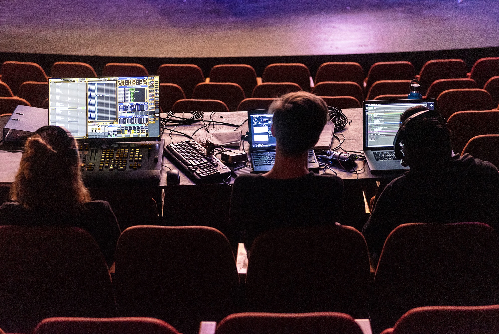 Three people sit at a control desk in a theatre, working on laptops and lighting consoles with multiple screens, facing an empty stage and rows of red theatre seats.