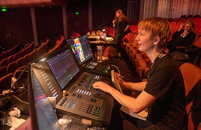 A person operates lighting and sound control desks in a theatre, with colourful screens and buttons visible. Another person stands nearby, and empty red seats fill the background.