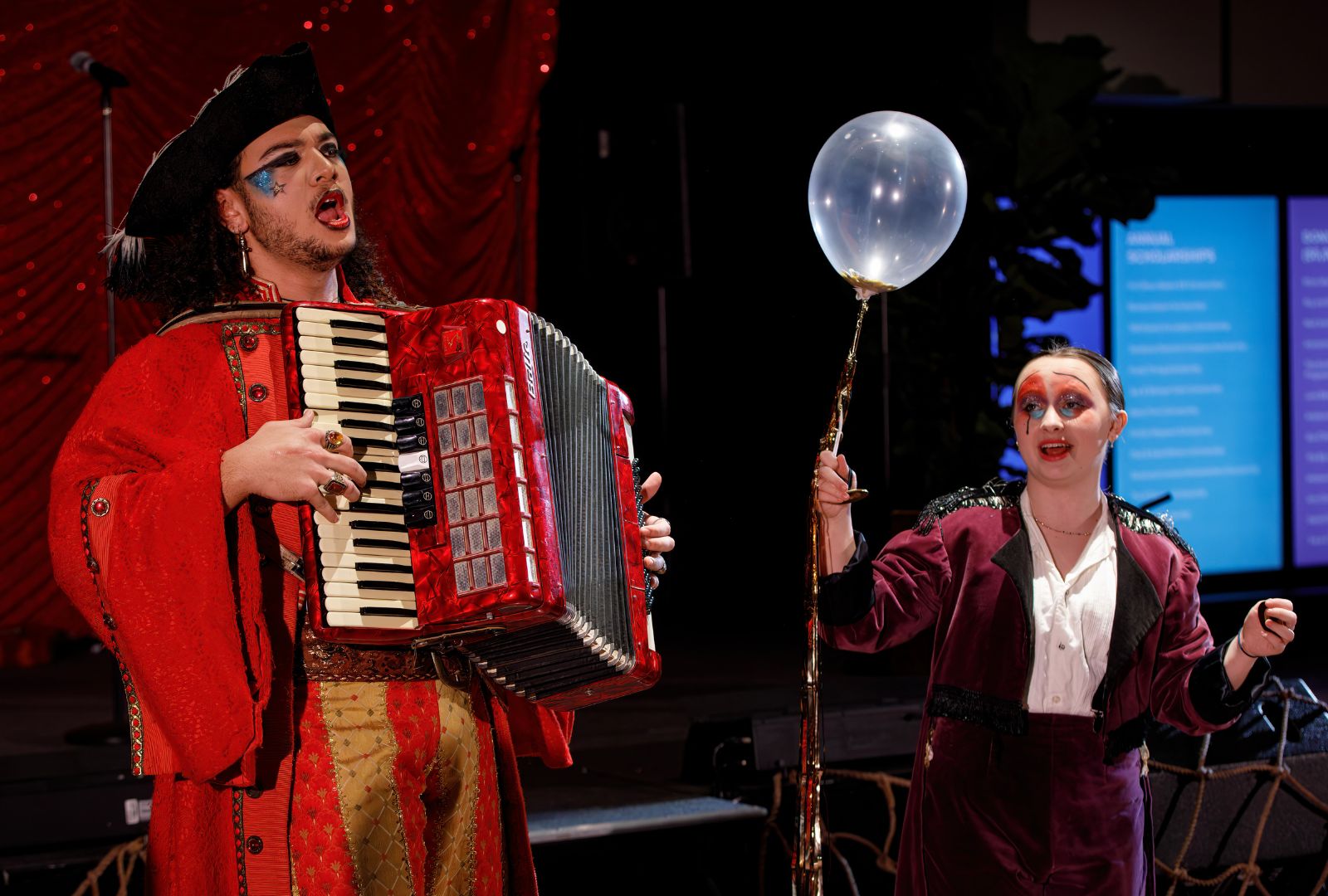 Tana Laga'aia (Musical Theatre, 2023) is dressed as a pirate in a red and gold coat, playing an accordion with Kayla Ingle-Olson (Musical Theatre, 2023) who is dressed in a purple suit and hold a balloon.