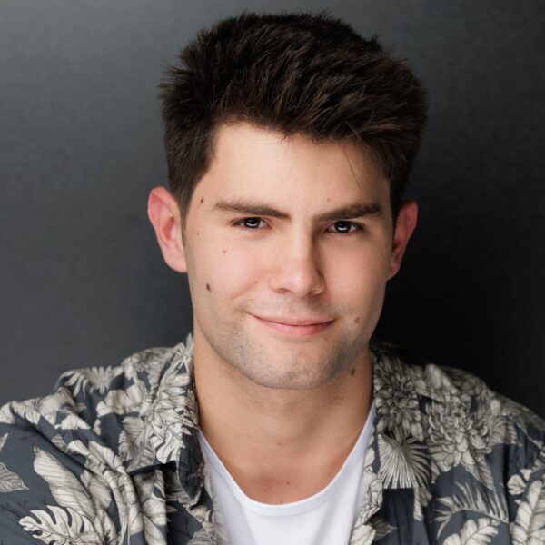 A young man with short dark hair smiles slightly at the camera. He is wearing a white T-shirt and a patterned shirt with floral designs, standing in front of a dark, plain background.