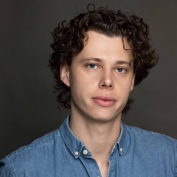 A young person with light skin, blue eyes, and curly brown hair is wearing a blue denim button-up shirt and looking directly at the camera against a dark, plain background.