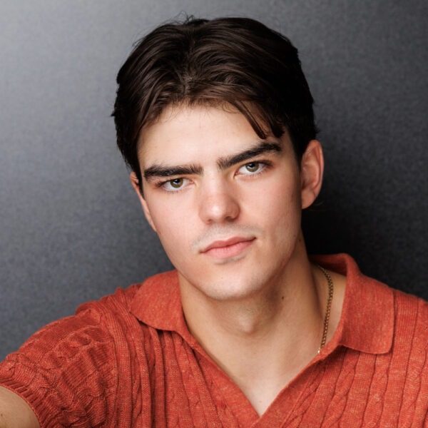 A young man with short dark hair and light skin wears a rust-orange textured polo shirt and looks directly at the camera with a neutral expression against a plain dark background.
