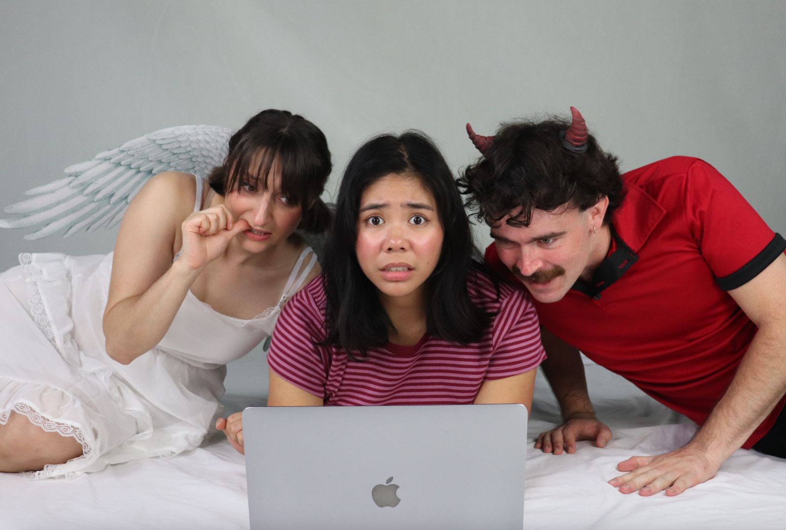 Three people are focused on a laptop. The centre person looks worried, flanked by a person in angel wings on the left and one with devil horns on the right.