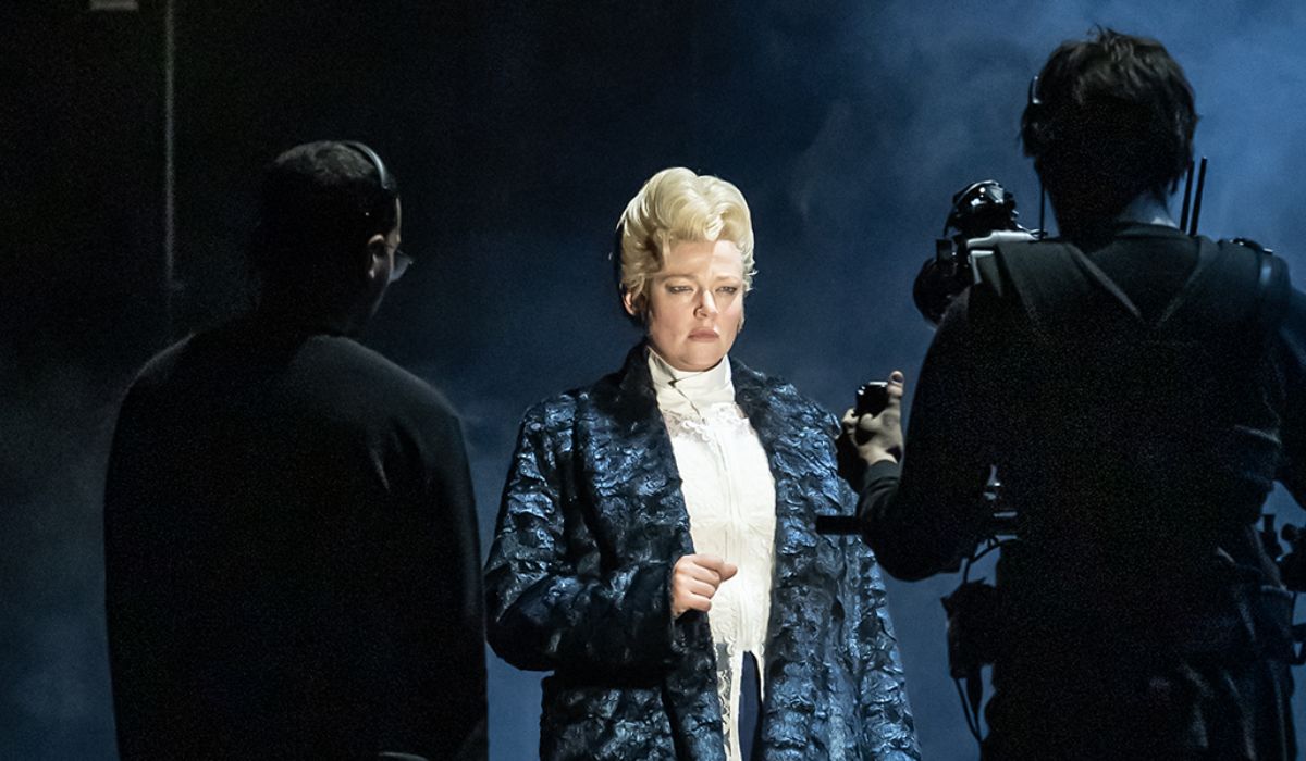 Sarah Snook as Dorian Gray in a blue coat with a blonde wig. There are two cameramen in the shadows facing her.
