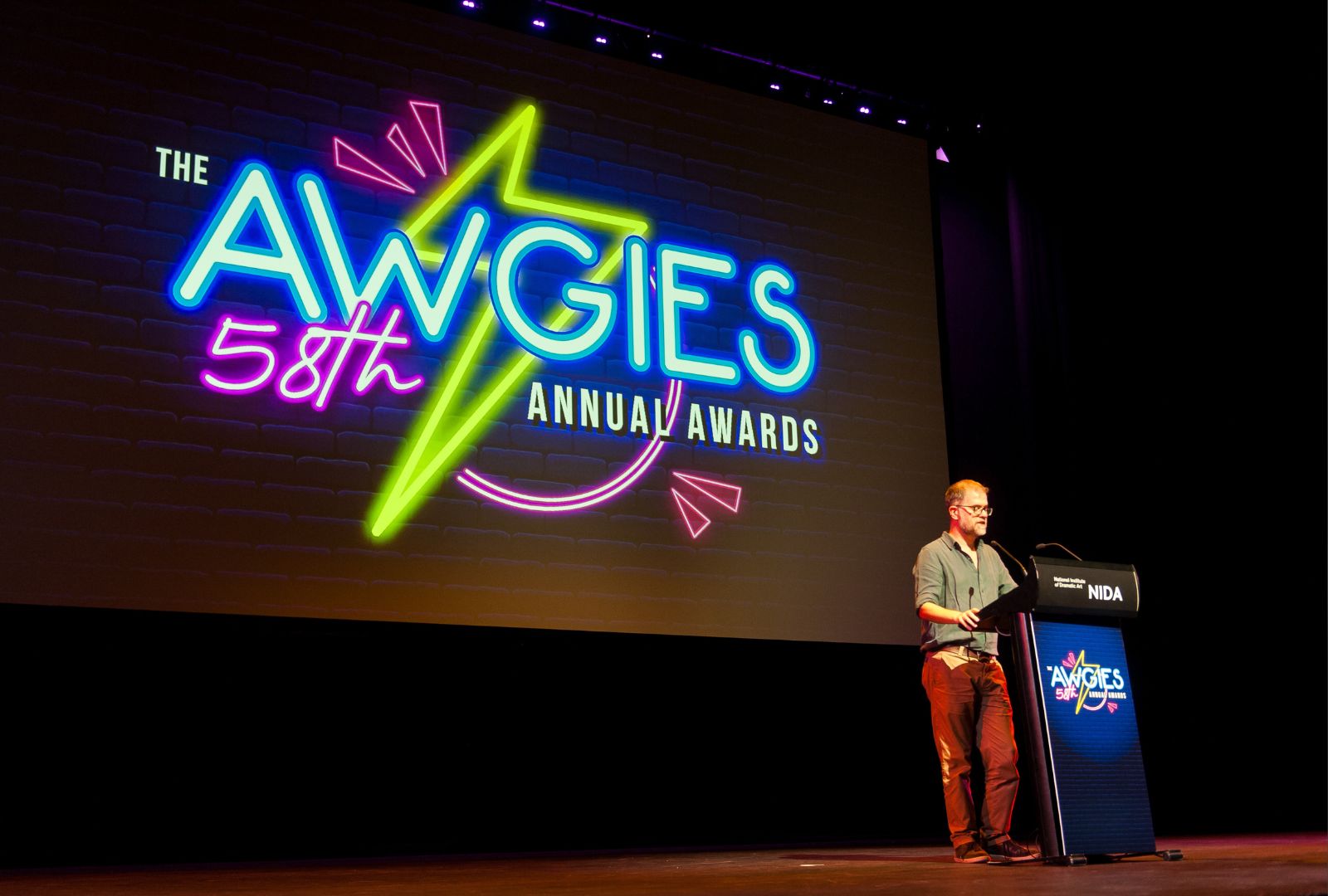 A person stands at a podium on a stage with a large screen displaying the text, "The AWGIES 58th Annual Awards." The podium has a logo for "AWGIES" and "NIDA."