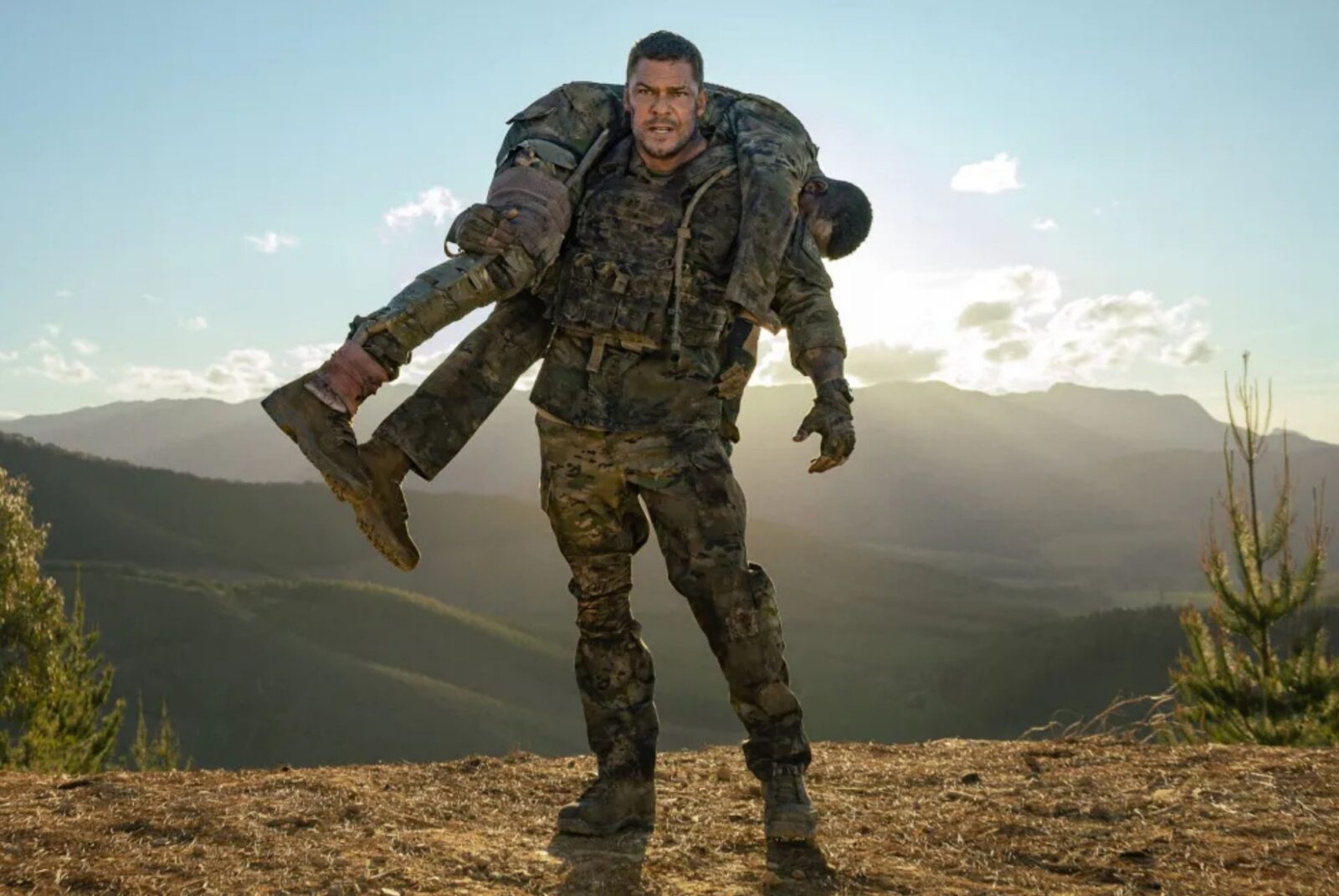 A soldier in camouflage gear carries another soldier over his shoulders with mountains and a bright sky in the background.