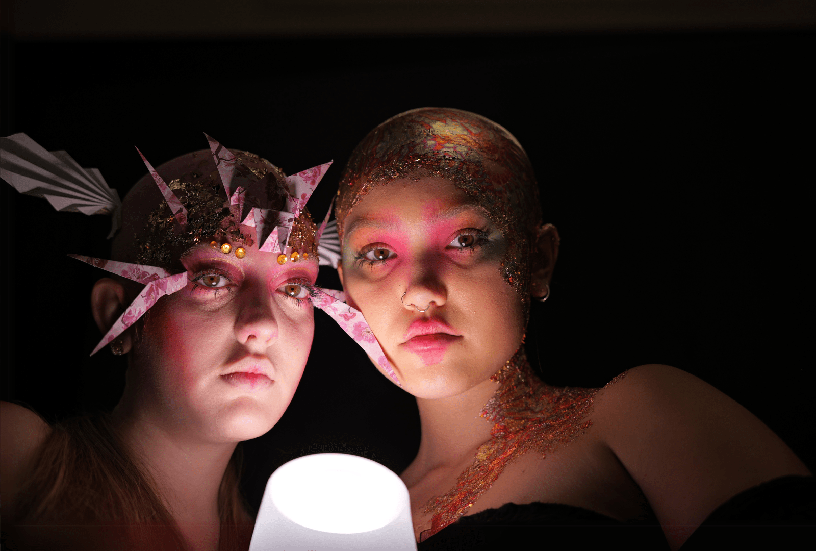 Two people in dramatic make-up and costumes stand close together in dim lighting, illuminated from below by a glowing white lamp. One wears a spiked headpiece; both have bold pink and glittery designs on their faces and heads.