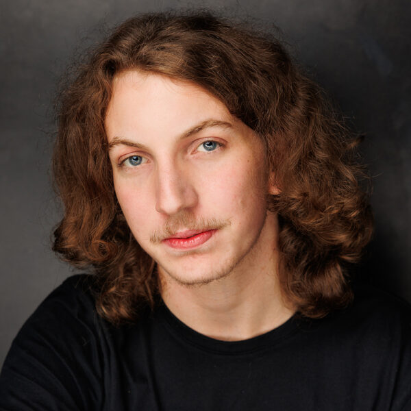 A young person with long, wavy brown hair, blue eyes, and a light moustache, wearing a black shirt, looks into the camera with a neutral expression against a dark background.