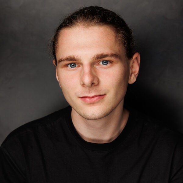 A young man with light skin, blue eyes, and long brown hair tied back, wearing a black shirt, looks at the camera with a slight smile against a dark background.